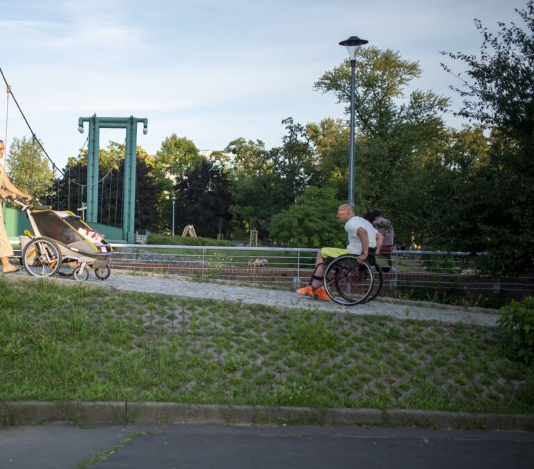 Zdjęcie wykonane na świeżym powietrzu latem. Na podjazd prowadzący do kładki dla pieszych wjeżdża Łukasz Siemież na wózku dla osoby z niepełnosprawnością. Naprzeciwko idzie kobieta z wózkiem dziecięcym, która dzięki podjazdowi może łatwo zjechać z kładki dla pieszych